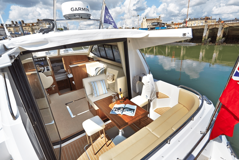 The cozy interior of a docked boat features plush cushioned seating and a polished wooden table adorned with an open magazine. Wine and glasses await as the helm and controls stand ready inside the inviting cabin.