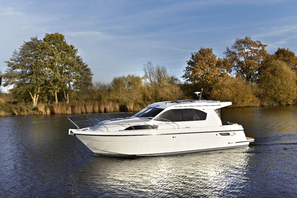 A white motor yacht glides gracefully on a calm river, surrounded by autumn trees under a clear blue sky—an idyllic scene where the pristine beauty of nature meets refined boat elegance.