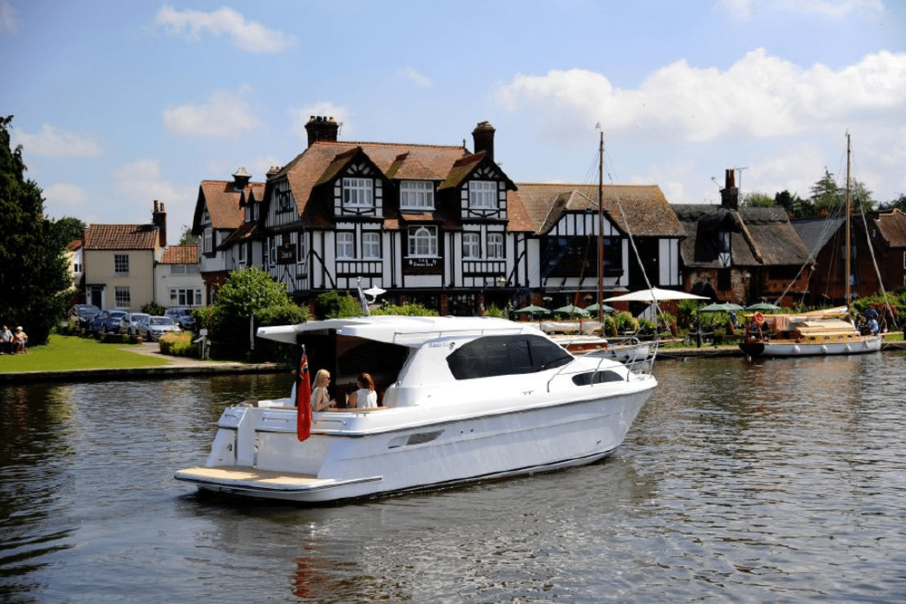 A white motorboat gracefully glides along the river, passing a charming, traditional-style house with a steep roof and black-and-white facade, under a partly cloudy sky.