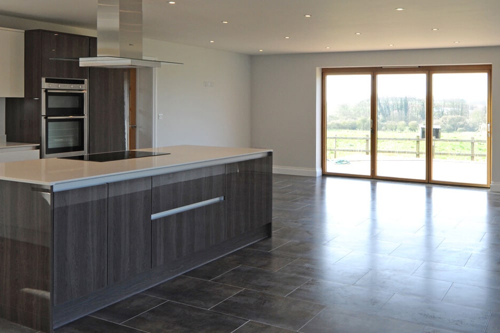 Modern kitchen with a large island and dark cabinetry, featuring tile flooring and a glass door leading to an outdoor view.