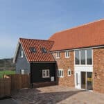 Thumbnail of http://Modern%20brick%20and%20wood%20house%20with%20red-tiled%20roof,%20large%20windows,%20and%20paved%20driveway,%20set%20against%20a%20rural%20landscape%20with%20a%20wooden%20fence%20and%20green%20fields.