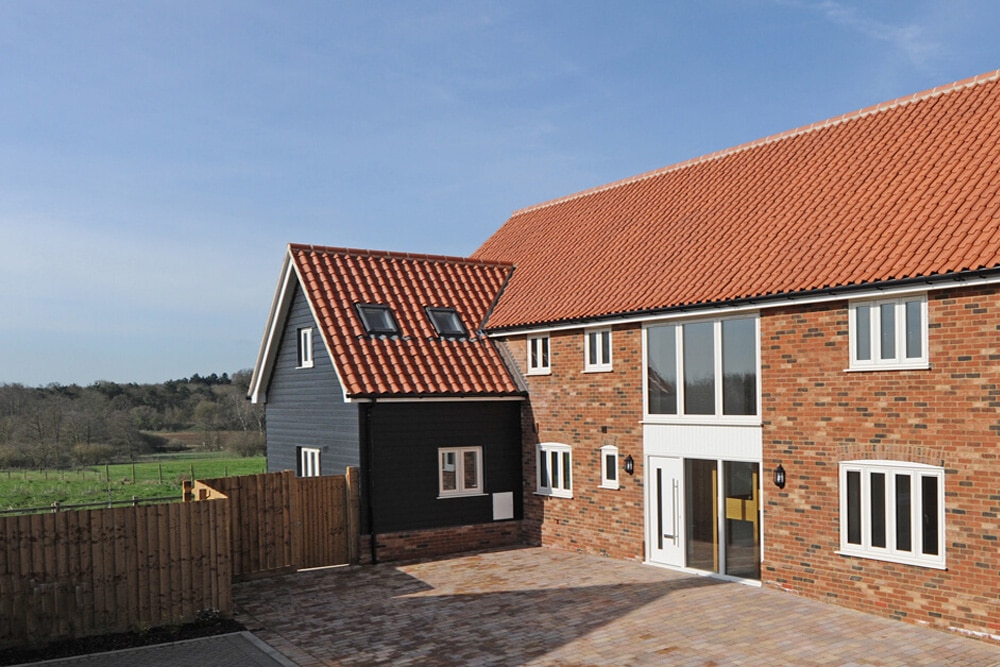 Modern brick and wood house with red-tiled roof, large windows, and paved driveway, set against a rural landscape with a wooden fence and green fields.