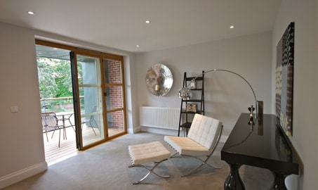 Modern living room with two white chairs, a curved floor lamp, a black table, metal wall art, and a shelving unit. Glass doors open to a balcony with outdoor seating.