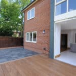 Thumbnail of http://Modern%20house%20with%20red%20brick%20exterior,%20large%20windows,%20and%20patio.%20The%20patio%20has%20a%20mix%20of%20wooden%20decking%20and%20stone%20tiles,%20adjacent%20to%20an%20open%20kitchen%20with%20gray%20cabinetry.