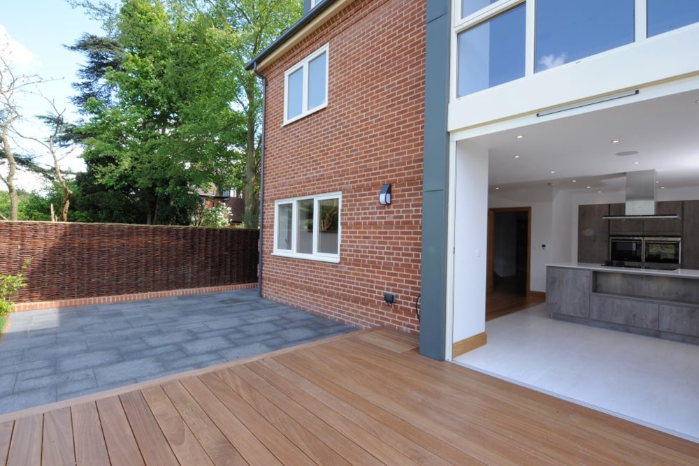 Modern house with red brick exterior, large windows, and patio. The patio has a mix of wooden decking and stone tiles, adjacent to an open kitchen with gray cabinetry.