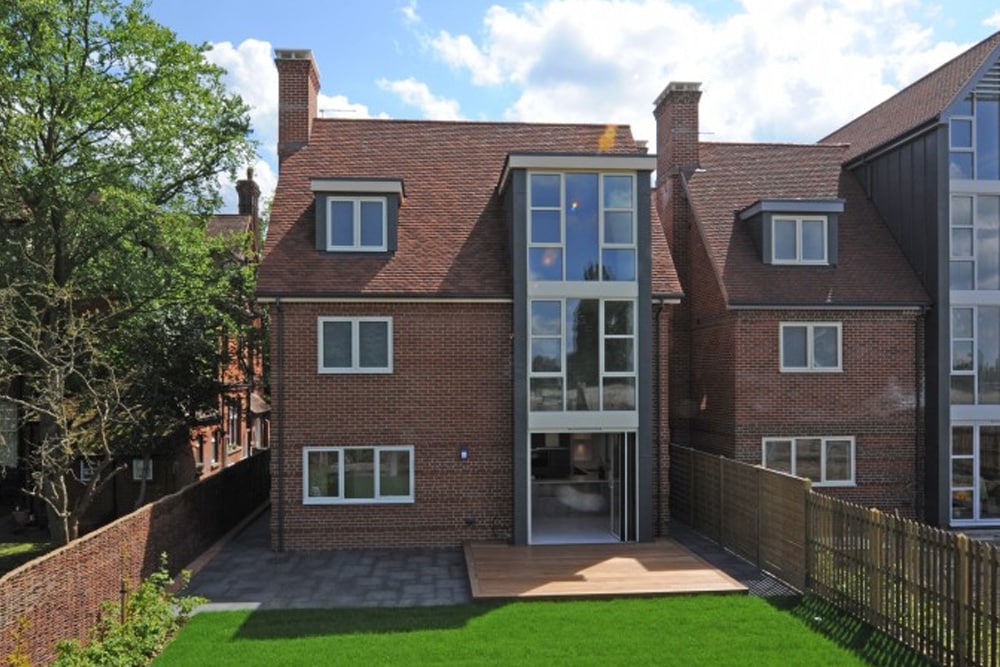 A modern red-brick house with large front windows, a small wooden deck, and fenced backyard with grass.