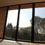 Thumbnail of http://Large%20glass%20sf75%20bifold%20doors%20open%20to%20a%20balcony%20with%20a%20view%20of%20trees%20and%20sky,%20framed%20by%20a%20wooden%20panel%20structure.%20Indoors,%20flowers%20are%20arranged%20by%20the%20entrance.