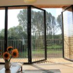 Thumbnail of http://Open%20glass%20bifold%20doors%20lead%20to%20a%20balcony%20with%20a%20view%20of%20trees.%20Inside,%20a%20wooden%20table%20with%20sunflowers%20and%20an%20orange%20sofa%20are%20visible.%20Sunlight%20streams%20in,%20illuminating%20the%20room.