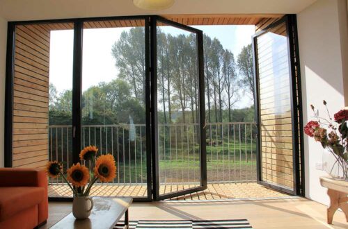 Open glass bifold doors lead to a balcony with a view of trees. Inside, a wooden table with sunflowers and an orange sofa are visible. Sunlight streams in, illuminating the room.