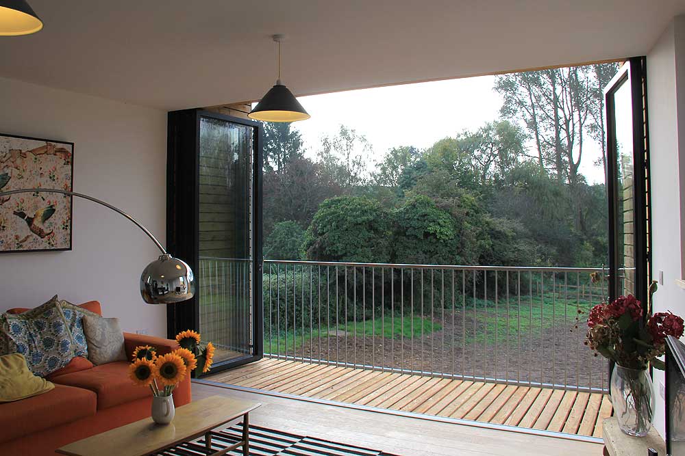The living room features an orange sofa, sunflowers on the table, and a view of a lush garden through sleek SF75 bifold sliding glass doors.