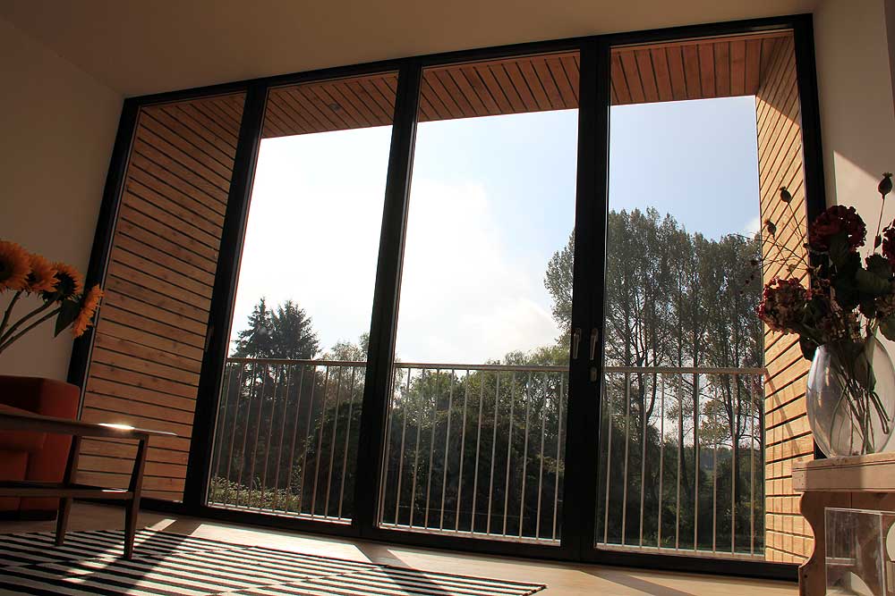 Large glass sf75 bifold doors open to a balcony with a view of trees and sky, framed by a wooden panel structure. Indoors, flowers are arranged by the entrance.