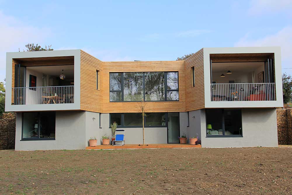 A modern two-story house with large windows and wooden accents, featuring SF75 bifold doors that open to two balconies, and several potted plants on the ground level.