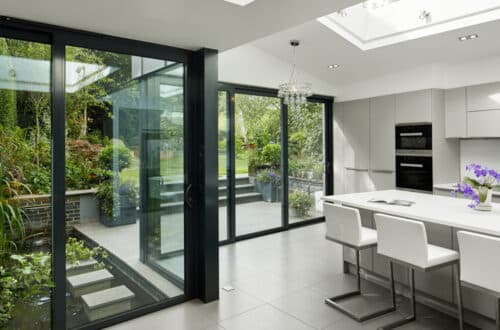 Modern kitchen with white cabinets and island, glass doors opening to a garden view. Skylight above, tiled floor, and barstool seating.