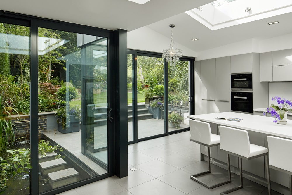 Modern kitchen with white cabinets and island, glass doors opening to a garden view. Skylight above, tiled floor, and barstool seating.