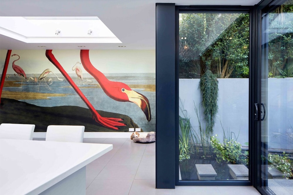 Modern room with large flamingo mural on the wall. Glass doors open to a small garden with minimal foliage. White furniture and sleek surfaces create a contemporary ambiance.