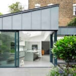 Thumbnail of http://Modern%20house%20with%20large%20glass%20sliding%20doors%20opening%20to%20a%20garden.%20A%20small%20dog%20sits%20on%20the%20floor%20inside%20the%20minimalist%20white%20kitchen.