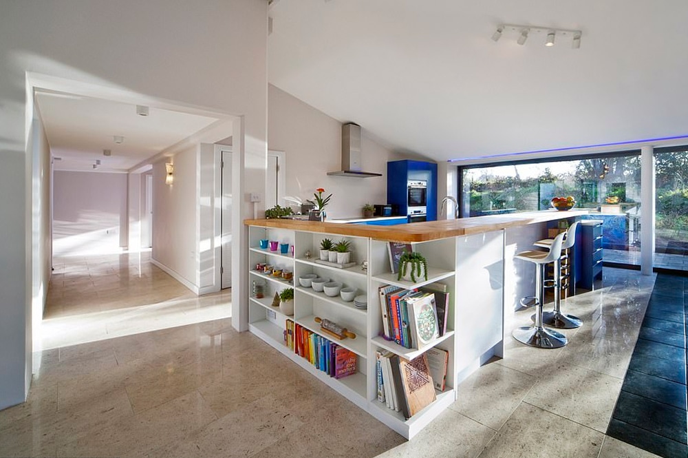 Modern kitchen with a central island, reminiscent of a Grand Designs feature, showcasing shelves of books and plants. It includes barstools and large windows offering garden views. Light tiled flooring and white walls enhance the bright space.