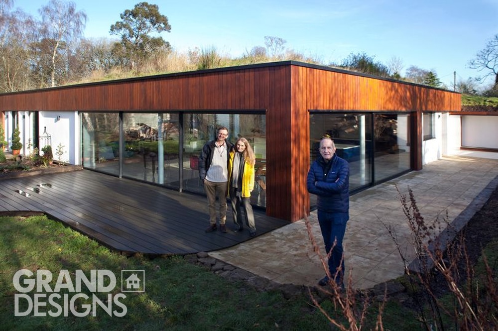 Two people stand by a striking, modern flat-roofed house with wooden siding and large windows, embodying the essence of "Grand Designs." Another person stands in the foreground with arms crossed, exuding confidence. The iconic "Grand Designs" logo adorns the bottom left corner.