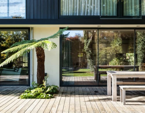 Modern house exterior with large glass doors, a wooden deck, a fern tree, and a wooden table. Reflections of greenery are visible in the windows.