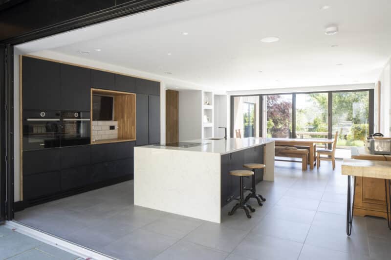 Modern kitchen with a large white island, black cabinetry, and a dining area. Floor-to-ceiling windows provide natural light. Two stools at the counter.