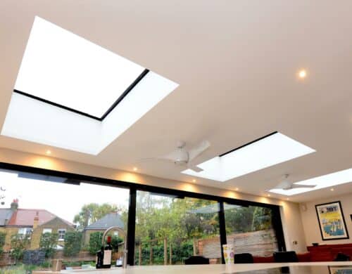 Modern interior with multiple flat rooflights featuring black frames and integrated ceiling lights, overlooking garden with traditional brick house.