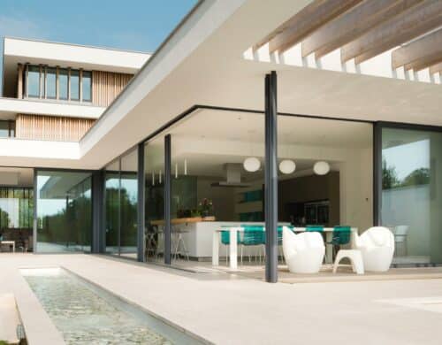 Modern house with large glass windows, a patio, and a narrow reflecting pool. The interior features a white and teal kitchen with contemporary decor.