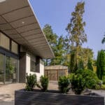 Thumbnail of http://Modern%20house%20exterior%20with%20floor-to-ceiling%20windows,%20large%20overhanging%20roof,%20and%20a%20patio.%20The%20scene%20includes%20a%20landscaped%20garden%20with%20various%20trees%20and%20shrubs%20under%20a%20clear%20sky.