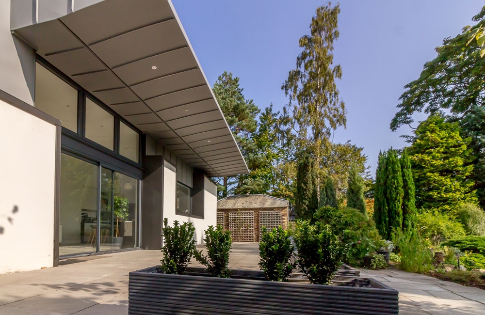 Modern house exterior with floor-to-ceiling windows, large overhanging roof, and a patio. The scene includes a landscaped garden with various trees and shrubs under a clear sky.