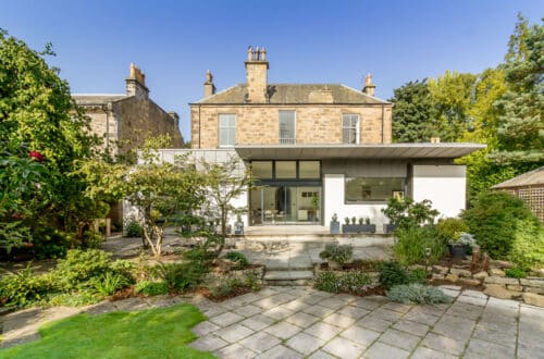 A two-story house with a mix of stone and modern white exteriors, surrounded by a garden with trees and shrubs, under a clear blue sky.