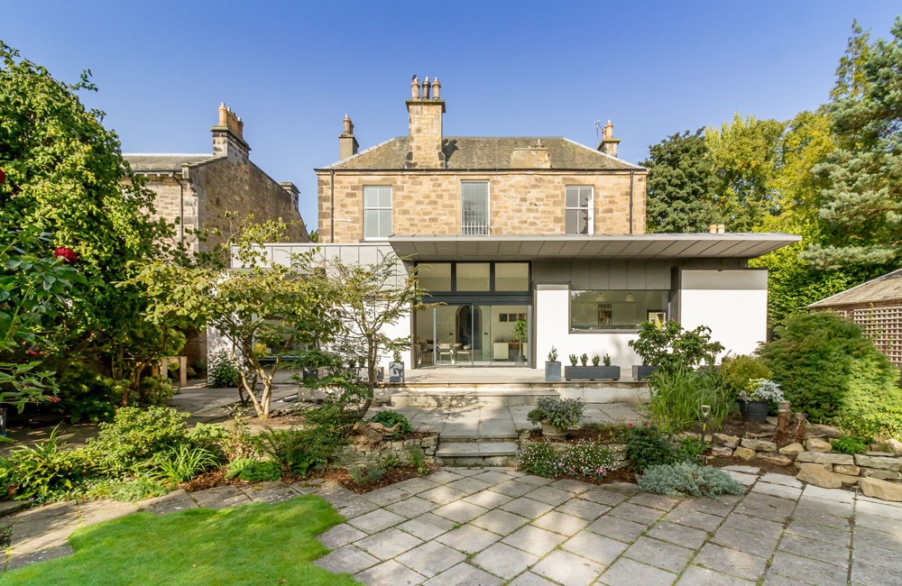 A two-story house with a mix of stone and modern white exteriors, surrounded by a garden with trees and shrubs, under a clear blue sky.