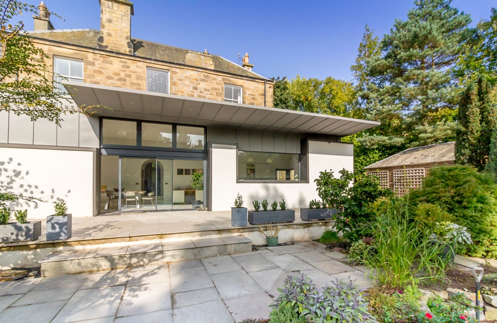 Modern extension on a stone house with large windows, a patio, and a landscaped garden featuring various plants and trees under a clear blue sky.