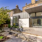 Thumbnail of http://Modern%20house%20exterior%20with%20large%20windows,%20a%20stone%20patio,%20and%20surrounding%20greenery.%20An%20older%20stone%20building%20is%20partially%20visible%20in%20the%20background%20under%20a%20clear%20blue%20sky.