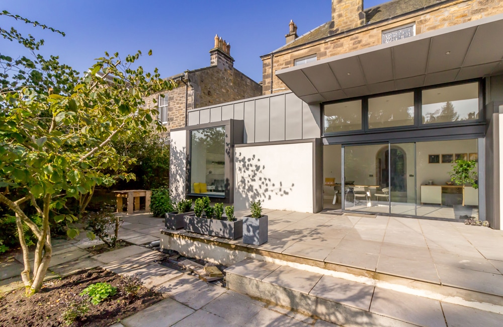 Modern house exterior with large windows, a stone patio, and surrounding greenery. An older stone building is partially visible in the background under a clear blue sky.