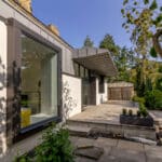 Thumbnail of http://Modern%20house%20with%20large%20windows%20and%20a%20flat%20roof,%20surrounded%20by%20trees%20and%20greenery.%20A%20patio%20area%20with%20concrete%20paving%20is%20visible%20in%20the%20foreground.