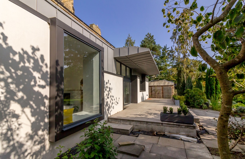 Modern house with large windows and a flat roof, surrounded by trees and greenery. A patio area with concrete paving is visible in the foreground.