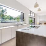 Thumbnail of http://Modern%20kitchen%20with%20large%20window,%20white%20cabinets,%20gray%20island%20with%20stove,%20breakfast%20bar,%20and%20pendant%20lights.%20Dining%20area%20visible%20in%20the%20background%20with%20greenery%20outside%20the%20window.