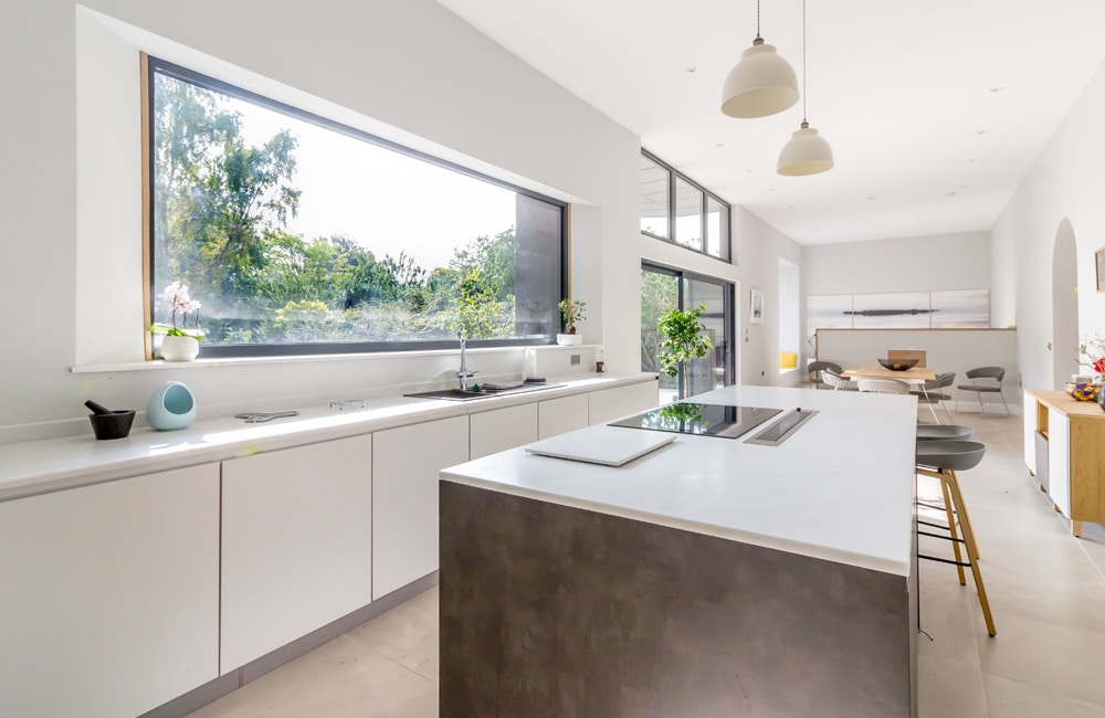 Modern kitchen with large window, white cabinets, gray island with stove, breakfast bar, and pendant lights. Dining area visible in the background with greenery outside the window.