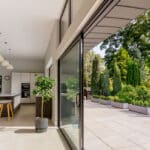 Thumbnail of http://Open-plan%20kitchen%20with%20a%20bar%20counter%20and%20stools,%20leading%20to%20a%20patio%20with%20potted%20plants%20and%20greenery%20visible%20through%20large%20sliding%20glass%20doors.