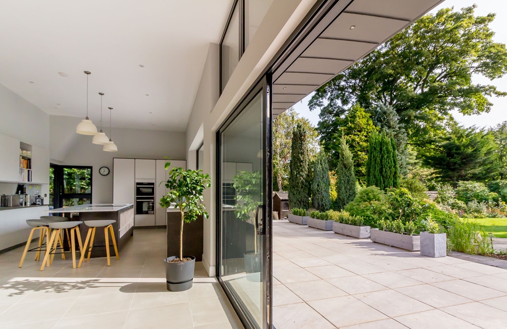 Open-plan kitchen with a bar counter and stools, leading to a patio with potted plants and greenery visible through large sliding glass doors.