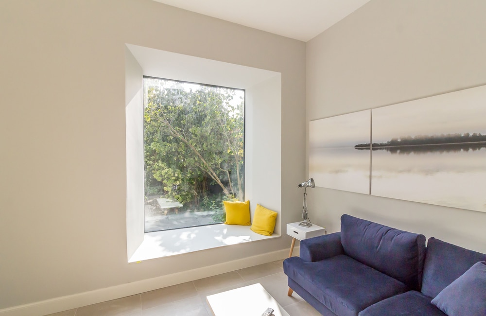 Minimalist room with large window, two yellow cushions, blue couch, white side table with a lamp, and serene wall art. Sunlight filters through the window, illuminating the space.