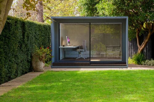 A small modern garden office with glass walls, featuring a desk, chair, and computer, is nestled amidst green hedges and a lush lawn.