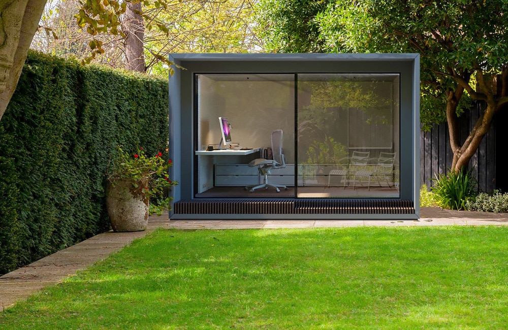 A small modern garden office with glass walls, featuring a desk, chair, and computer, is nestled amidst green hedges and a lush lawn.