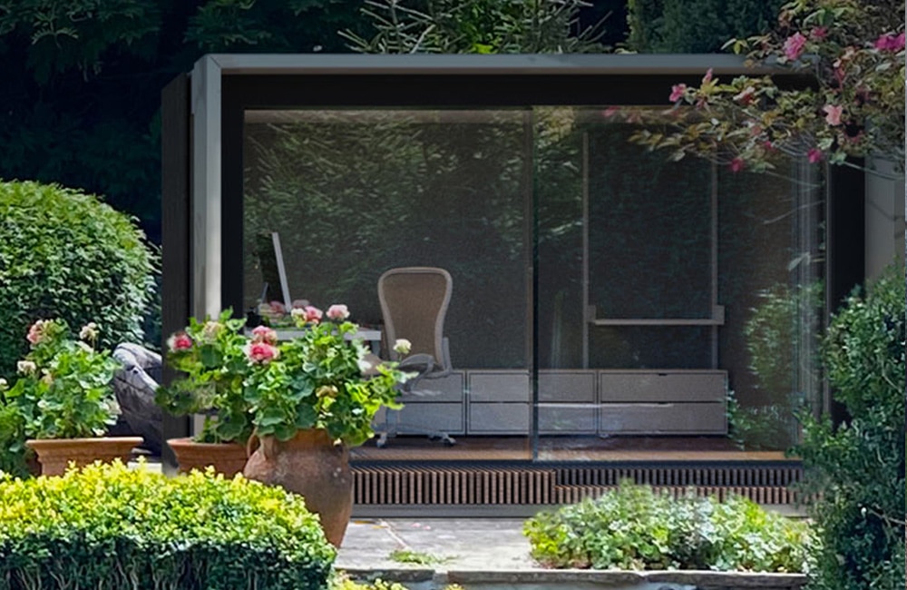 A modern glass garden office with a white chair and a desk, surrounded by lush green plants and colorful flowers.