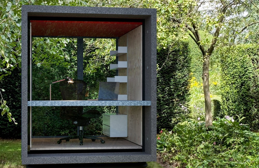 A small, modern garden office cube with glass walls, a desk, chair, and shelves, surrounded by greenery.