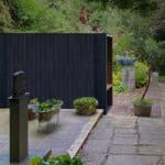 Thumbnail of http://A%20modern%20black%20structure%20stands%20in%20a%20garden%20with%20stone%20paths,%20surrounded%20by%20green%20foliage%20and%20potted%20plants.