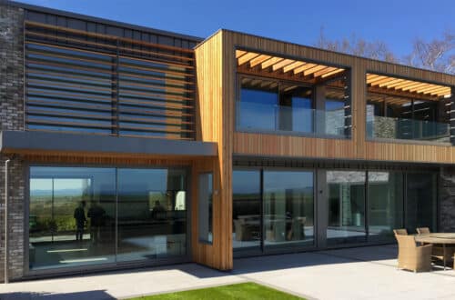 Modern house exterior with large glass windows, brick and wood paneling. A patio with outdoor furniture is visible in the foreground under a clear blue sky.