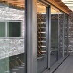 Thumbnail of http://Modern%20building%20exterior%20with%20large%20glass%20windows%20and%20brick%20walls.%20The%20covered%20walkway%20features%20a%20wooden%20lattice%20roof%20casting%20shadows%20on%20the%20passage.