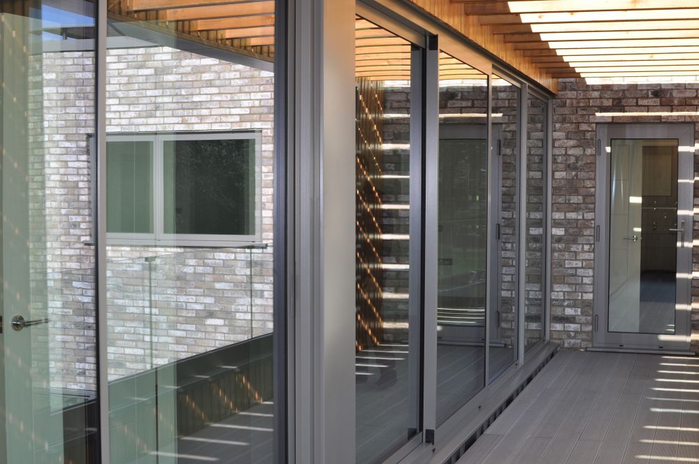 Modern building exterior with large glass windows and brick walls. The covered walkway features a wooden lattice roof casting shadows on the passage.