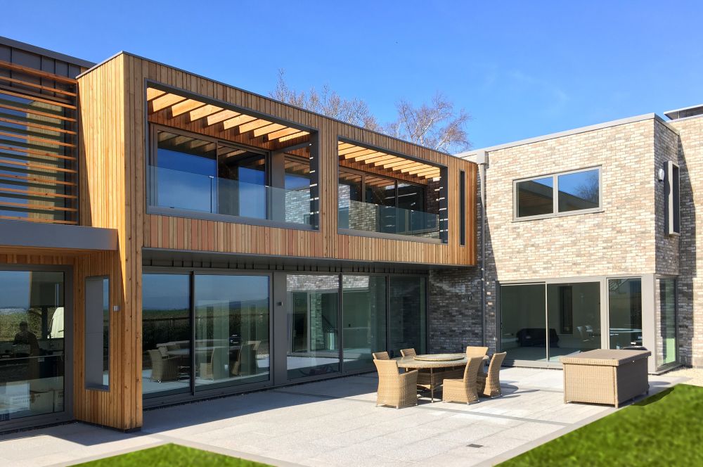 Modern house with wooden and brick exterior, featuring large windows, a balcony, and a patio with outdoor furniture set.