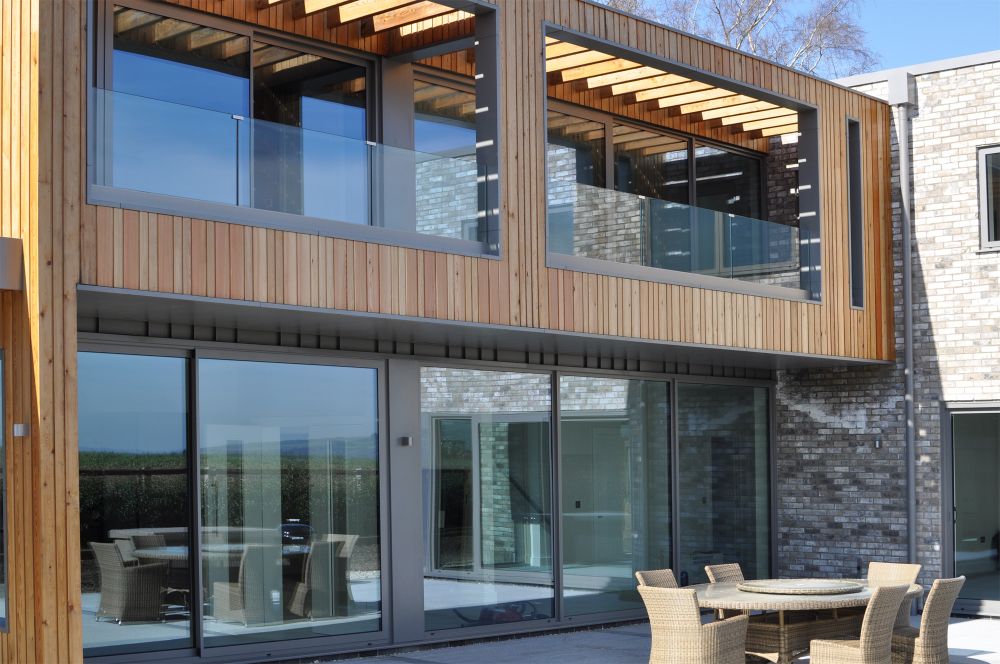 Modern house exterior with large glass windows, wooden paneling, and a brick facade. Outdoor patio set with a round table and chairs in the foreground.